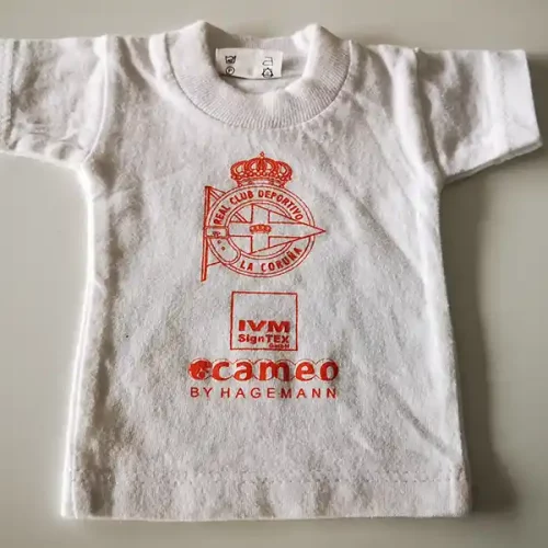 Un t-shirt blanc pour bébé avec le logo rouge du R.C. Deportivo La Coruña bien en évidence au centre, avec une couronne et une bande diagonale. En dessous, en texte rouge, on peut lire « IVM SignTex » et « cameo BY HAGEMANN », mettant en valeur lexpertise en matière de personnalisation textile.