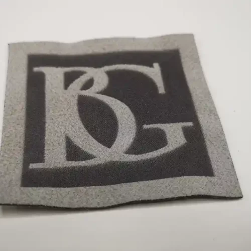 Un patch en tissu gris et noir présente de grandes lettres entrelacées « B » et « G » dans une police stylisée, parfaite pour la personnalisation textile. Les lettres blanches se détachent sur le fond sombre, encadré par une bordure grise. Le patch repose élégamment sur une surface blanche unie.