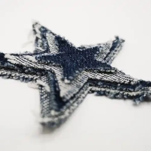 Un gros plan dun patch en denim effiloché en forme détoile, présentant une texture superposée et diverses nuances de bleu, incarne le charme du textile personnalisé. Le patch repose élégamment sur une surface blanche.