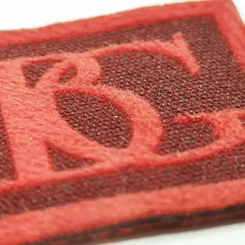 Gros plan dun patch texturé mettant en valeur les lettres rouges entrelacées « B » et « G » sur un fond carré rouge plus foncé, parfait pour les projets textiles de personnalisation.
