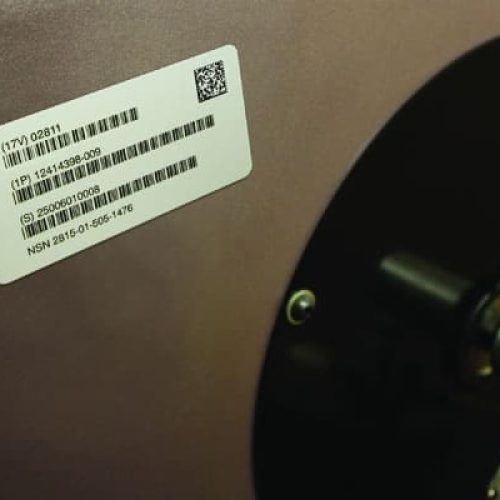Gros plan d'une étiquette de code-barres blanche avec un texte noir et un code QR attachés à une surface métallique près d'un composant mécanique circulaire noir et or.