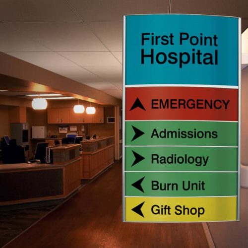 Intérieur d'un hôpital avec un panneau indiquant les directions pour les urgences, les admissions, la radiologie, l'unité des brûlés et la boutique de cadeaux ; la réception et les ordinateurs sont visibles à l'arrière-plan à côté d'une machine laser double source.