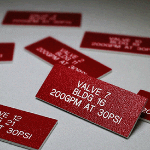 Plusieurs étiquettes en plastique rouge, fabriquées à partir de matière à graver en fraisage avec un texte gravé en blanc comme "VALVE 7 BLDG 16 200GPM AT 30PSI", sont disposées sur une surface blanche, certains détails étant nets et d'autres légèrement flous.