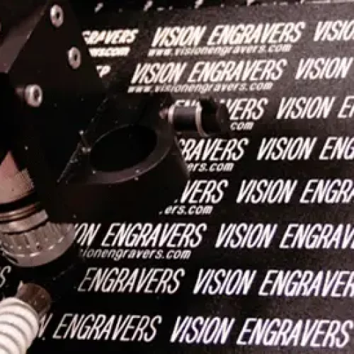 Gros plan dune machine à graver en action, gravant à plusieurs reprises « Vision Engravers » et lURL « visionengravers.com » sur une surface noire. Les composants de la machine, notamment une lentille et un bras, sont visibles.