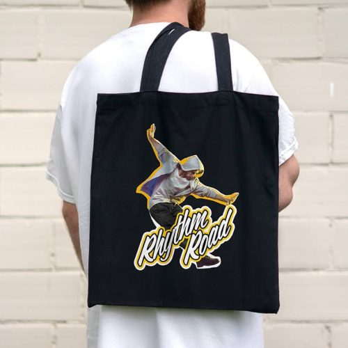 Une personne portant une chemise blanche tient un sac fourre-tout noir, probablement fabriqué avec une imprimante à jet dencre, avec un motif représentant une silhouette dansante portant un sweat à capuche et le texte « Rhythm Road ». Larrière-plan est un mur de briques claires.