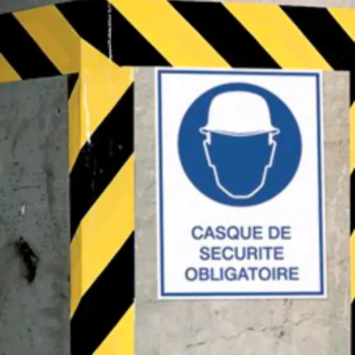 Un chantier de construction avec un panneau représentant un casque de sécurité blanc à lintérieur dun cercle bleu, indiquant « Casque de sécurité obligatoire ». Des bandes de danger noires et jaunes entourent le panneau.