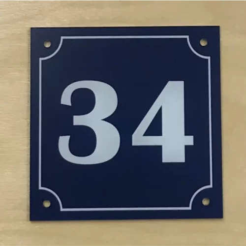 Un panneau carré fixé au mur affiche le numéro « 34 » en blanc sur fond bleu foncé, avec des bordures décoratives et des coins arrondis. Quatre petits trous sont situés près des coins du panneau.