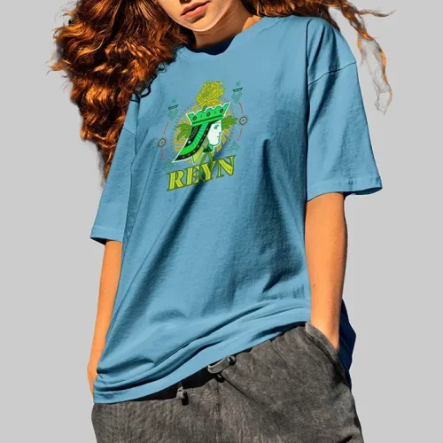 Une personne aux longs cheveux roux ondulés porte un t-shirt bleu ample avec un graphisme vert et le mot REYN, créé à l'aide de l'impression DTF, ainsi qu'un pantalon gris foncé, sur un fond gris uni.