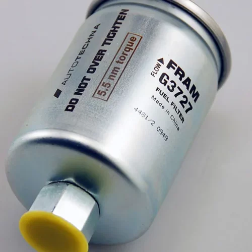 Un filtre à carburant cylindrique en métal étiqueté "FRAM G3721" est couché sur le côté, avec un texte clair marqué au laser CO2 : "NE PAS TROP SERRER", "Couple de 5,5 Nm" et "FILTRE À CARBURANT". L'extrémité du connecteur est munie d'un capuchon jaune.