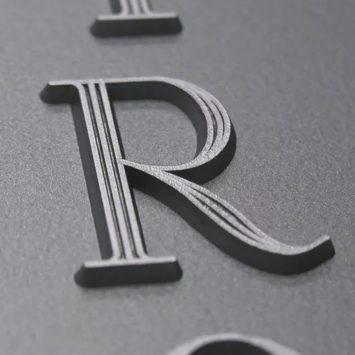 Gros plan d'une lettre R en relief et en relief avec des lignes parallèles gravées sur une surface lisse, gris foncé. L'image met l'accent sur la texture et les détails de la lettre.