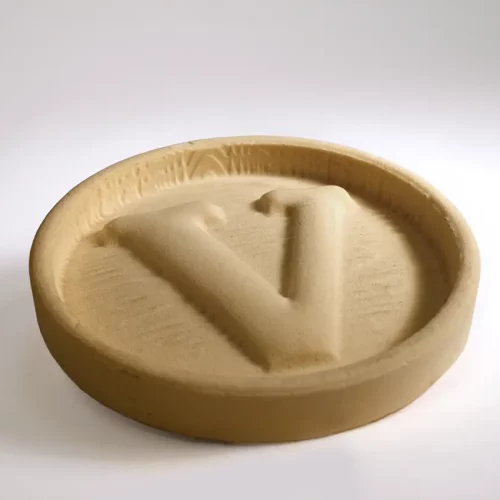 Un plateau rond de couleur beige avec la lettre V en relief au centre, fabriqué dans un matériau qui ressemble à du sable ou à de la poudre comprimée, placé sur une surface blanche unie.