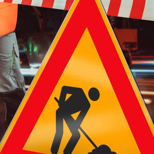 Un panneau d'avertissement triangulaire jaune et rouge pour la construction, probablement fabriqué avec un traceur de découpe, montre une silhouette noire creusant devant une barrière rayée rouge et blanche. Une partie d'un ouvrier vêtu d'un gilet orange est visible à gauche.