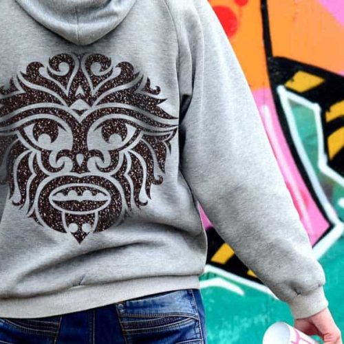 Une personne portant un sweat à capuche gris avec un motif de masque tribal dans le dos se tient près dun mur de graffitis vibrant, une bombe aérosol et un traceur de découpe à la main, créant de lart abstrait avec des tons roses, orange et verts.