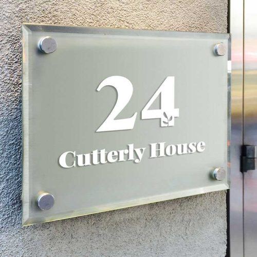 Une enseigne en verre montée sur un mur gris affiche le numéro « 24 » et le nom « Cutterly House » en texte blanc, réalisés de main de maître avec un plotter de découpe. Lenseigne est fixée par des entretoises métalliques élégantes à chaque coin.