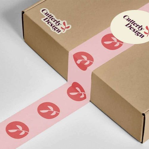 Une boîte en carton fermée par du ruban adhésif rose avec des illustrations de cerises rouges est présentée, ce qui laisse deviner la précision de sa fabrication. La boîte porte un autocollant « Cuttery Design », mettant en valeur le nom de la marque, probablement créé à laide dun plotter de découpe pour des détails complexes.