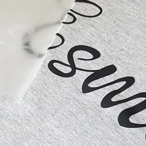 Un gros plan sur une surface grise révèle un lettrage en vinyle noir, minutieusement réalisé à laide dun plotter de découpe, partiellement recouvert dune feuille de transfert transparente. Le mot commence par « sm » dans une élégante police cursive, le reste étant masqué par la feuille.