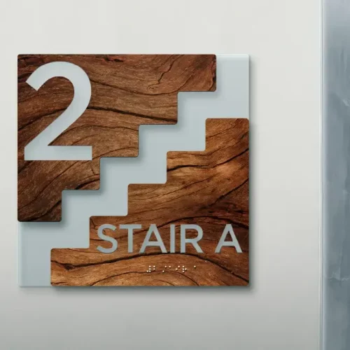 Un panneau sur un mur indique "2 STAIR A" en grosses lettres avec du braille en dessous. Réalisée en bois et en verre, l'enseigne - avec son graphisme d'escalier - a été imprimée sur objet pour indiquer l'escalier du deuxième étage.