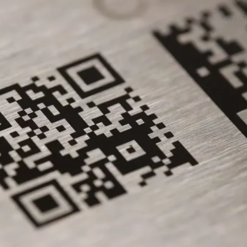 Vue rapprochée d'un code QR noir et blanc gravé au laser marqueur sur une surface métallique brossée. Un autre code QR partiel, créé à l'aide de la technologie laser co2, est visible dans le coin supérieur droit de l'image.