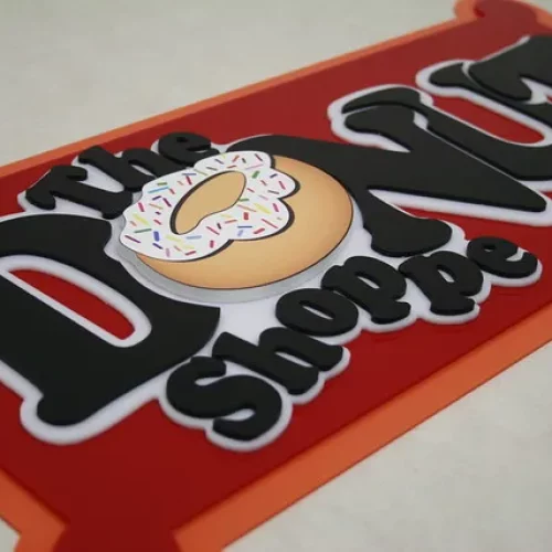 Une enseigne indique The Donut Shoppe avec un donut illustré comme la lettre O de Donut. Le beignet, recouvert d'un glaçage blanc et de paillettes colorées, suggère la précision des machines de fraisage en héliogravure. L'enseigne a un fond rouge et une bordure orange.