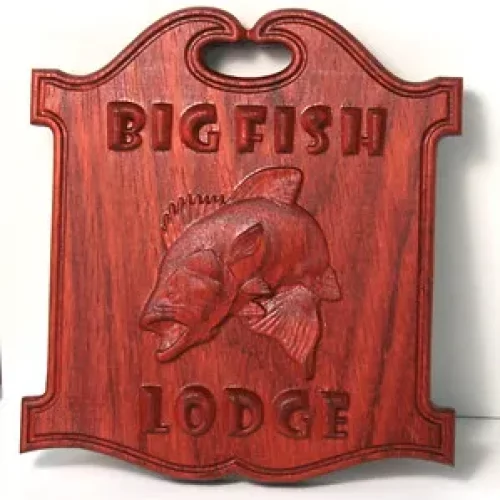 Une plaque en bois fabriquée à l'aide d'une défonceuse CNC Vision présente un poisson gravé au centre et les mots BIG FISH LODGE sont gravés au-dessus et en dessous. Elle présente une finition brun-rouge et comporte une petite poignée découpée au sommet.