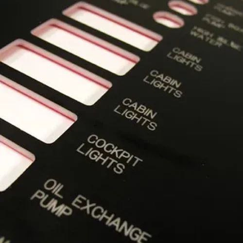 Vue rapprochée d'un panneau de commande noir, probablement issu de machines de fraisage en héliogravure, avec des découpes rectangulaires étiquetées CABIN LIGHTS, COCKPIT LIGHTS, et OIL EXCHANGE PUMP en texte blanc.