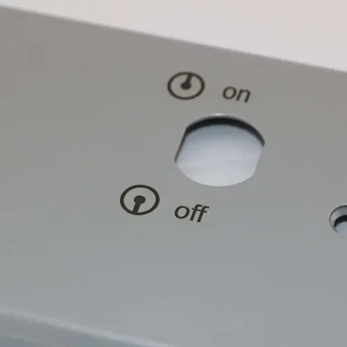 Gros plan d'un panneau métallique gris pour un marqueur laser, avec un trou circulaire étiqueté "on" en haut et "off" en bas, chacun avec des symboles de puissance. Le panneau comporte également d'autres découpes et un petit logo dans le coin inférieur gauche.