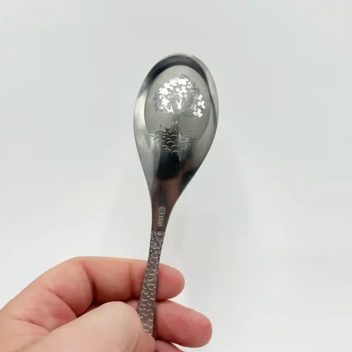 Une main tient une cuillère en métal finement gravée dun motif darbre sur le bol, sur fond blanc uni. Cette belle pièce met en valeur lart de graver le métal, mettant en valeur un savoir-faire exquis dans chaque détail délicat.