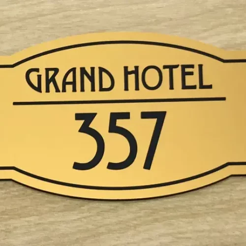 Une plaque de chambre dhôtel de couleur dorée avec un texte noir indiquant « Grand Hotel » au-dessus du numéro « 357 » sur un fond en bois.