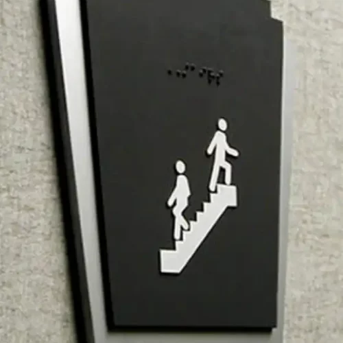 Panneau tactile et en braille avec des personnages descendant des escaliers, indiquant un escalier ou une cage descalier. Le panneau a un design moderne avec des couleurs contrastées contre le mur.