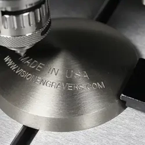 Une machine à graver en métal grave les mots MADE IN USA WWW.INSIDEMGRAVERS.COM sur une plaque ronde fixée par des équerres, mettant en valeur le travail des experts sur la matière à graver en fraisage.