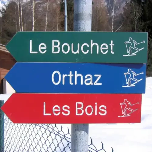Un panneau indicateur dans un paysage enneigé indique Le Bouchet (vert), Orthaz (bleu) et Les Bois (rouge), chacun avec des icônes de skieurs. Une cabane en bois et des arbres sont visibles en arrière-plan.