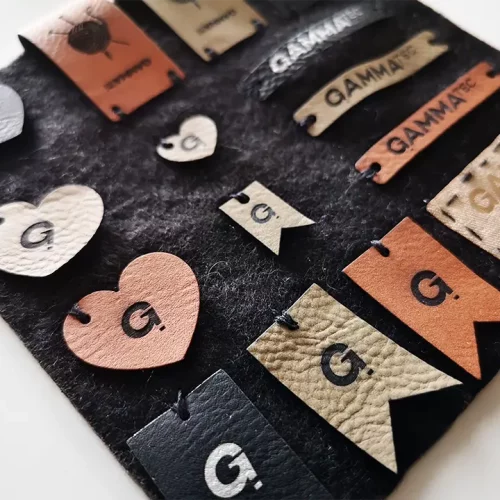 Une collection d'étiquettes en cuir et en tissu, comprenant des cœurs, des rectangles et des drapeaux, tous attachés à un fond de feutre noir. La plupart des étiquettes comportent la lettre G. et le mot GAMMA dans différentes polices.
