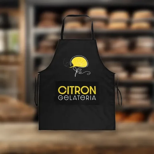 Un tablier noir comporte une illustration de tranches de citron jaune et le texte CITRON GELATERIA en jaune et blanc. L'arrière-plan montre des étagères avec du pain ou des pâtisseries floues.