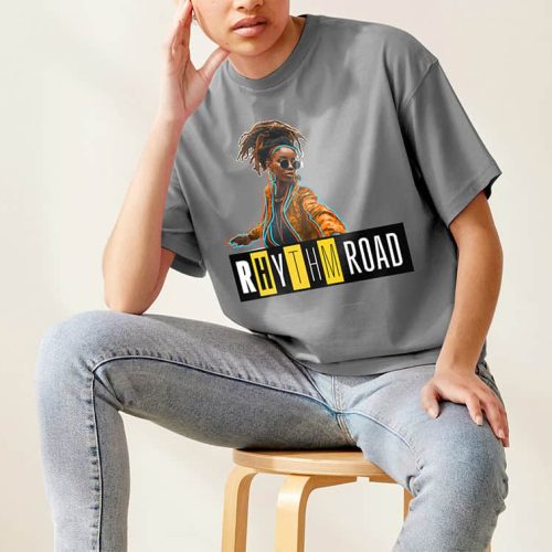 Une personne assise sur un tabouret porte un t-shirt gris avec un imprimé à jet dencre représentant une femme élégante et le texte « Rhythm Road », associé à un jean bleu clair. La personne pose son coude sur son genou, dans une pose détendue sur un fond uni.