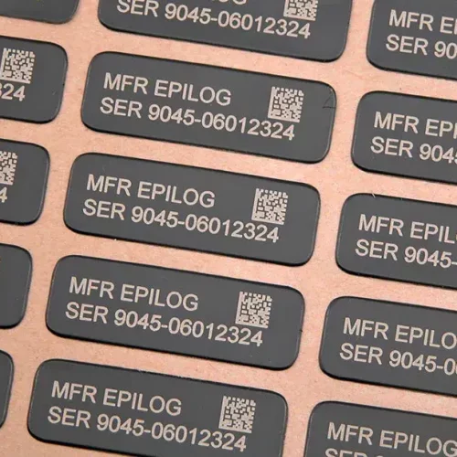 Vue rapprochée de plusieurs étiquettes rectangulaires noires créées à l'aide d'un laser graveur, comportant un texte imprimé en blanc indiquant MFR EPILOG SER 9045-06012324 et un code QR sur chacune d'entre elles, disposées en rangées ordonnées sur une surface plane.